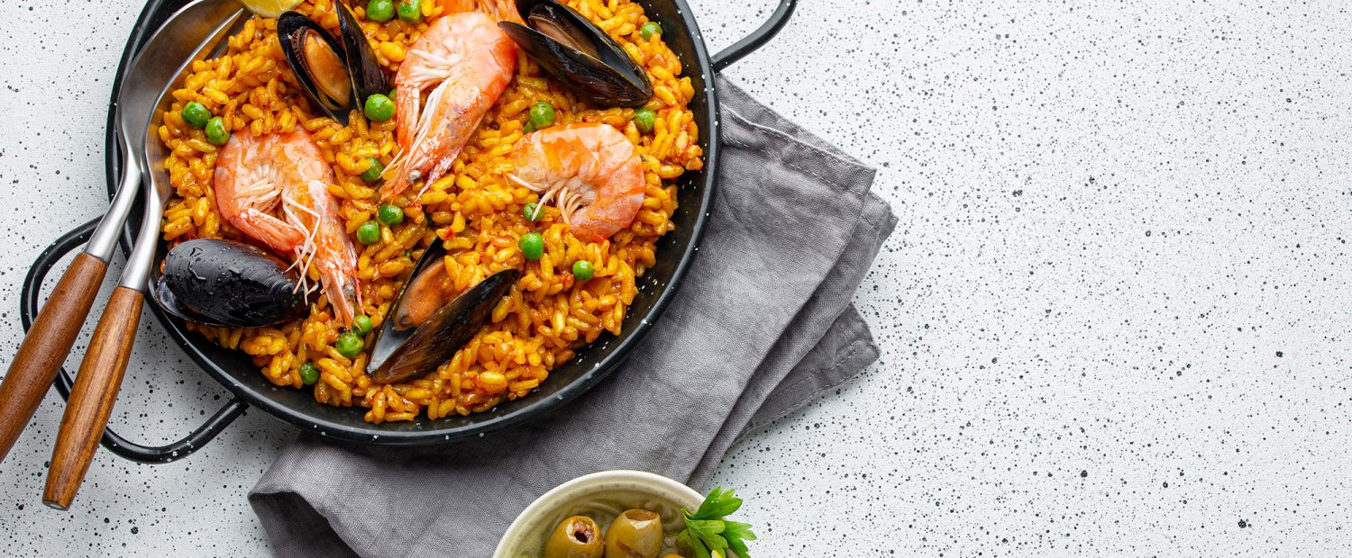 Classic dish of Spain, seafood paella in traditional pan on white wooden background top view. Spanish paella with shrimps, clamps, mussels, green peas, fresh lemon wedges from above, space for text